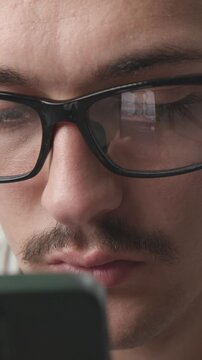 Close-up of a young man wearing glasses focused on his smartphone, screen reflecting in his eyeglasses 