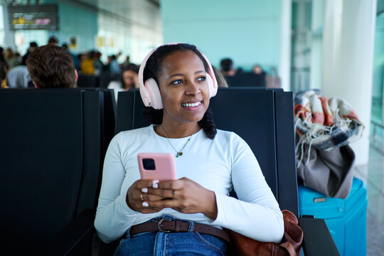 Young woman using phone to be connected and enjoying music on headphones, waiting for flight at a modern airport.
