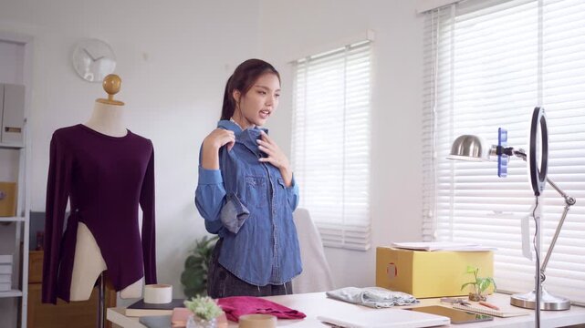 Young Asian online seller presenting blue denim shirt to camera while live streaming product from home studio
