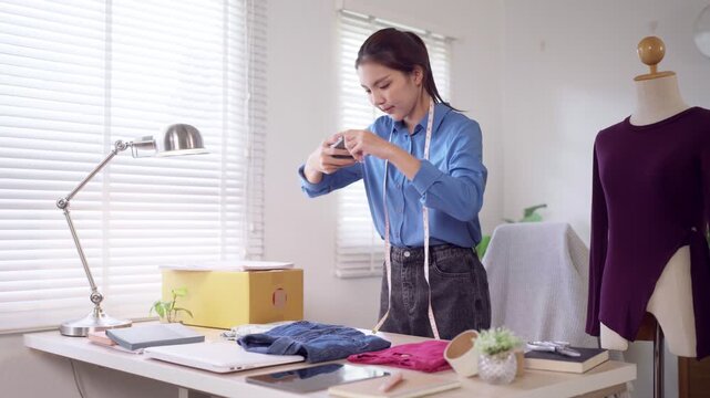 Young Asian fashion seller photographing clothing merchandise with smartphone while managing online boutique at home workspace
