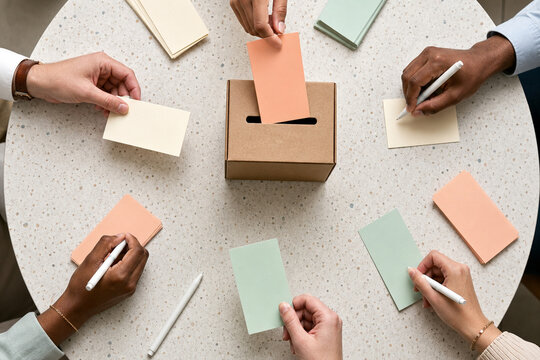 Overhead view of diverse hands writing on colorful ballot cards and placing a vote into a cardboard box during a group decision.