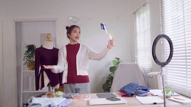 Young Asian fashion seller presenting clothing options to camera while recording product video in bright home studio