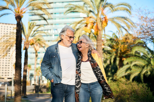 Smiling senior couple walking arm in arm through a sunny park, sharing laughter and affection. Casual denim style, palm trees in the background, relaxed stroll on a bright day.