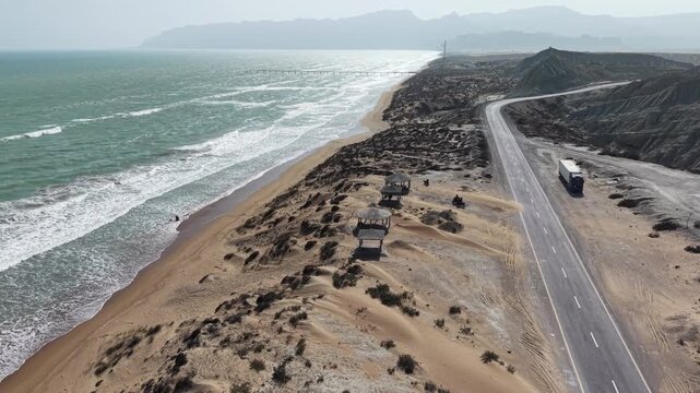 Drone view of the Makran Coastal Highway in Balochistan, Pakistan, featuring a scenic Arabian Sea coastline, rugged mountains, sandy beaches, winding road, and breathtaking coastal landscape.