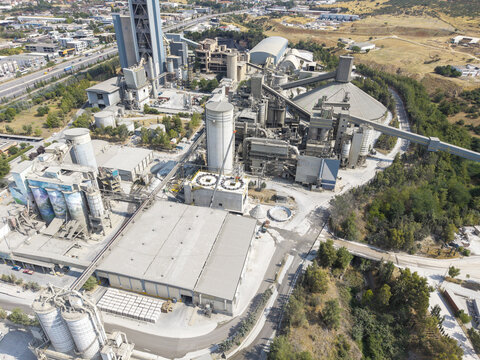 Aerial view of TITAN Cement factory featuring industrial silos, conveyor belts, and large storage facilities surrounded by trees and roads in Thessaloniki, Central Macedonia, Greece.