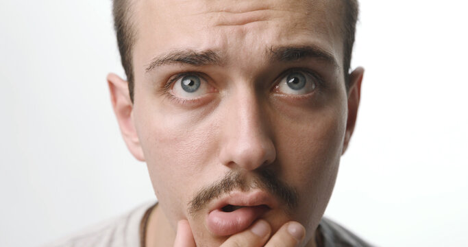 Confused and pensive young man with mustache and blue eyes touching chin on white studio background.
