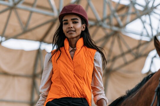 Young female equestrian in orange riding vest and helmet portrait on horseback in covered arena for lifestyle banner and equestrian sport design