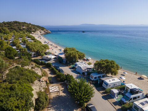 Aerial view of a beachside campsite in Armenistis featuring rows of caravans parked under lush trees along the turquoise waters of the Aegean Sea, Armenistis, Greece.
