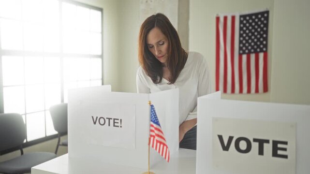 Woman places ballot into a white voting booth at a polling table in building with american flag and privacy screen visible; civic duty pride.