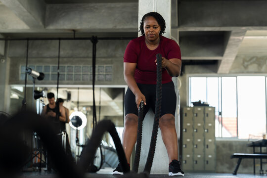 Plus size African American woman training with battle rope waves in modern gym, wide fitness lifestyle photo for cardio strength workout, endurance routine, healthy living and body confidence.