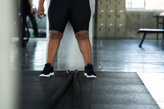 Close up of plus size African American woman standing by battle rope in modern gym, fitness lifestyle photo for cardio strength workout preparation, active wellness, endurance and body confidence.