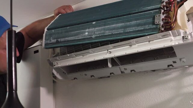 Technician lifting front panel of wall mounted split air conditioner for cleaning service, exposing evaporator coil, copper tubing, indoor housing and repair details