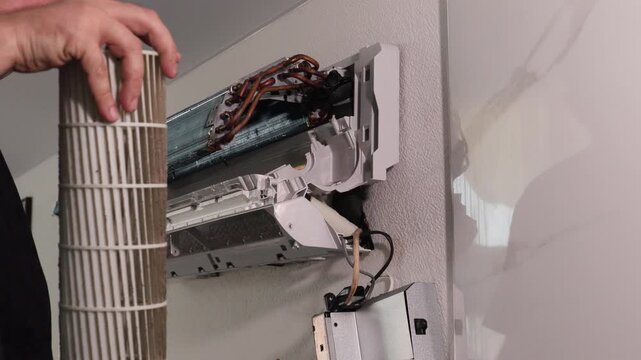 Technician removing evaporator coil and blower wheel from wall mounted split air conditioning unit during indoor service maintenance with exposed wiring, brackets, and metal housing