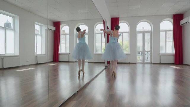 Elegant ballerina in a blue tutu and pointe shoes gracefully rehearsing classical ballet moves, including jumps and pirouettes, in a spacious, sunlit dance studio with a large mirror