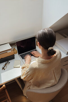 Back view of woman working on laptop at a sunny home office desk.