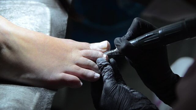 	
Close-up of a professional nail technician using an electric nail drill with a ceramic bit to remove old gel polish from a client's toenail during a pedicure. Professional foot care.