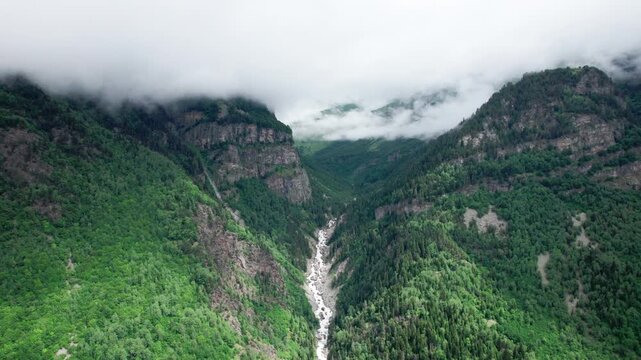 Aerial drone flight over a lush green mountain valley with a crystal-clear river flowing through the gorge. Towering peaks, pristine summer wilderness and breathtaking alpine scenery in the Caucasus