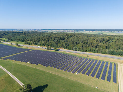 Aerial view of a large solar farm with rows of photovoltaic panels set in a green field near a forest and highway in Germany.