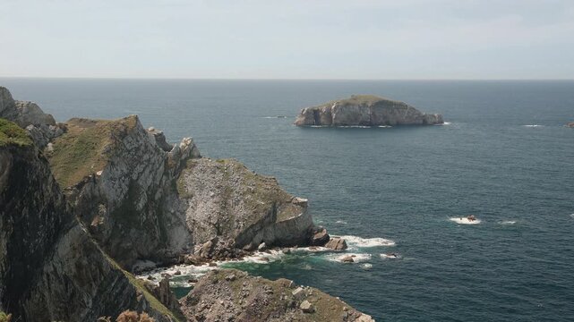 Scenic footage of the rocky coastline at Cape Pe&ntilde;as in Asturias, Spain, showing steep cliffs, crashing waves, and a serene islet on a beautiful summer day