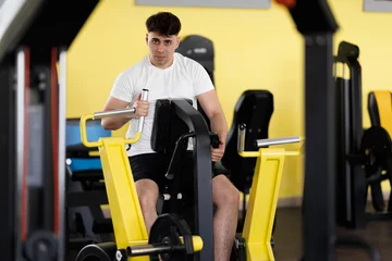 Fotobehang Sportschool Young man works out on gym equipment in fitness center during day focusing on upper body strength and fitness training  © Nikola Spasenoski