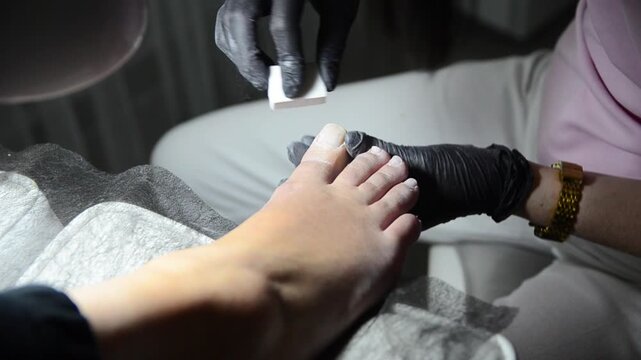 Professional pedicure master smoothing and buffing toenails with a nail buffer during a salon treatment. Close-up of foot care procedure preparing nails for the next stage of pedicure.