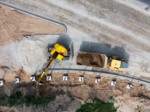 Aerial view of a construction site with an excavator loading soil into a dump truck near concrete foundation pillars in Kaunas, Kaunas County, Lithuania.