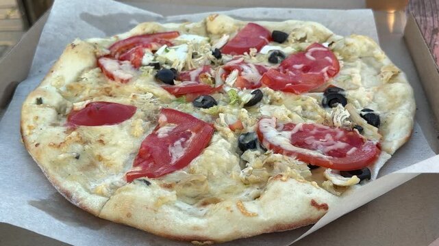 Baked pizza with tomato slices, black olives, chicken and cheese in a cardboard box. Fast food delivery, italian cuisine, takeaway meal