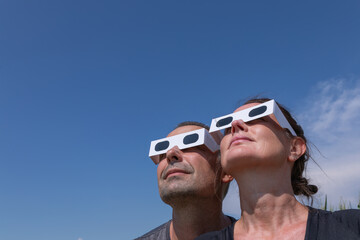 Low-angle close-up view of a man and a woman wearing protective paper glasses, looking up into a clear blue sky to observe the upcoming solar eclipse on August 12, 2026.