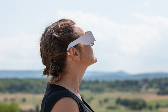 Horizontal side profile portrait of a woman wearing protective solar eclipse glasses looking up toward the sky over a scenic rural countryside landscape.