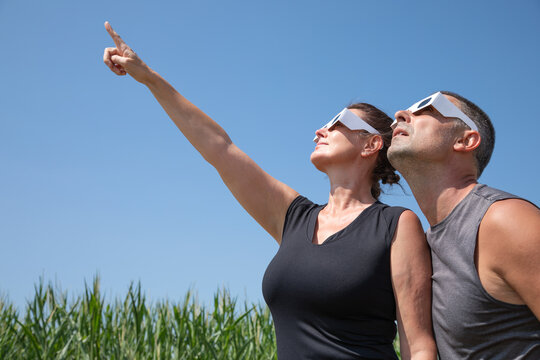 Medium close-up profile view of a woman and a man wearing protective paper glasses, looking up into the sky to observe the upcoming solar eclipse on August 12, 2026.