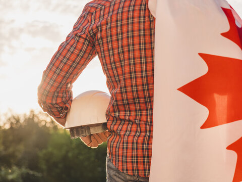Construction worker holds a white hard hat with a Canadian flag draped over his shoulder. Symbolizes Canadian workers or Labor Day. Concept of pride in construction profession