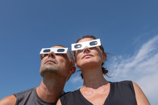  man and a woman wearing protective paper glasses, with the bright sunburst reflecting in the lenses as they look up into a clear blue sky to observe the upcoming solar eclipse on August 12, 2026.