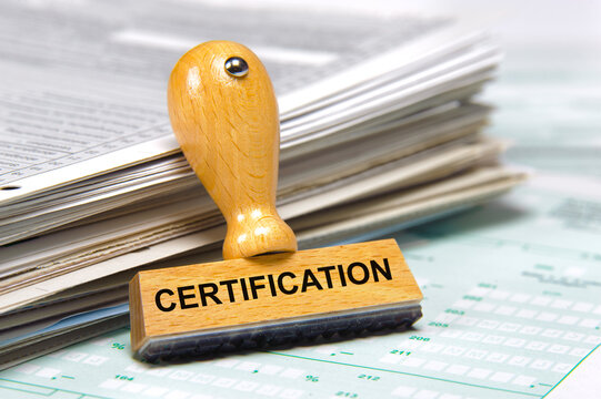 In einem B&uuml;ro lehnt ein Holzstempel an einem Stapel Papiere mit der Aufschrift: certification