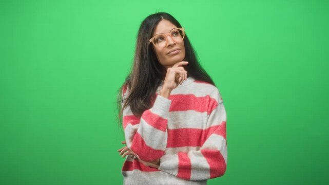 Woman with hand on chin and glasses wearing a striped sweater and looking pensive against a green studio backdrop; thoughtful introspection.