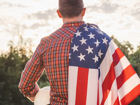Construction worker holds a white hard hat with an American flag draped over his shoulder. Symbolizes American workers or Labor Day. Concept of pride in construction profession and American dream