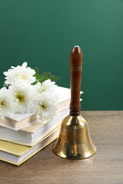 Golden school bell, flowers and stack of books on wooden table near blackboard, closeup