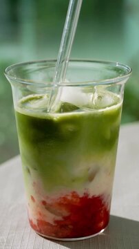 A detailed close-up shot of a glass cup with iced strawberry matcha latte. A transparent glass straw slowly stirs the layers, creating beautiful green, white, and red swirling patterns