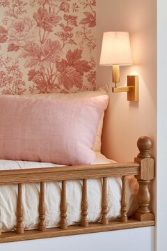 Close-up of a built-in loft bed nook featuring cream cotton bedding and a pink decorative pillow against a floral wallpaper backdrop with a wall-mounted lamp