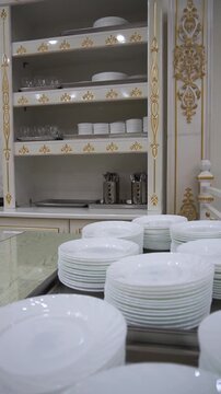 Slow motion camera panning of stacks of clean white ceramic plates on a table with a luxury white and gold classic cupboard in the background, vertical video.