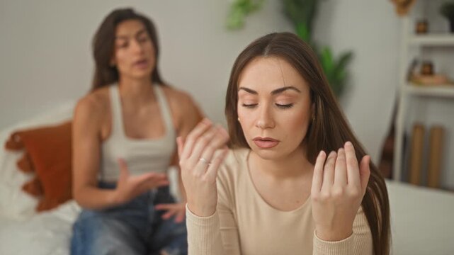 Woman sitting on bed touching temples with hands while a friend speaks behind her in a bedroom apartment setting; frustration support.