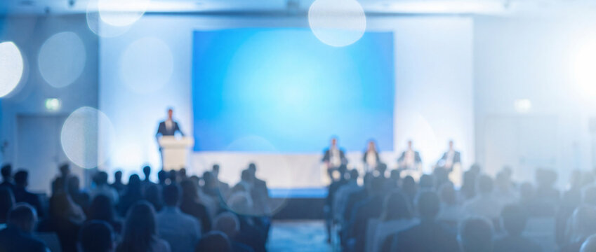 Business conference hall with blurred audience and speaker on stage featuring bright blue screen display, ideal for corporate event presentation backdrop or website banner