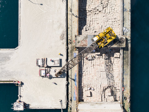 Aerial view of Celukan Bawang Port showing a yellow crane loading cement onto a cargo ship with workers on the dock in Kecamatan Gerokgak, Bali, Indonesia.