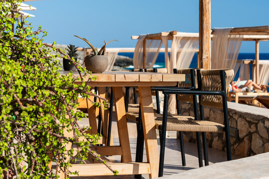 Close up of a beach bar setting in Greece, showcasing a wooden table with woven chairs and succulents, on a terrace with cabanas and the striking turquoise sea backdrop perfectly completing the scener