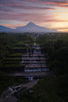Aerial view of Grojogan Watu Purbo, a multi-tiered waterfall with cascading water, lush vegetation, and Mount Merapi at sunset in Kecamatan Tempel, Daerah Istimewa Yogya, Indonesia.
