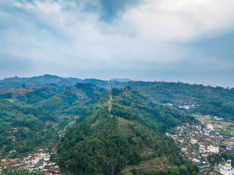 Aerial view of a telecommunications tower standing on a lush green hill overlooking a rural village with dense vegetation and rolling hills in Pasirjambu, West Java, Indonesia.
