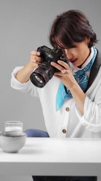 A woman in her thirties is using a camera to capture images while sitting at a table in a bright studio space that shows a simple background.
