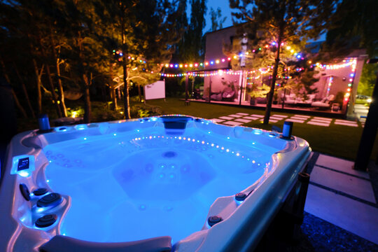 Warm water jacuzzi sits in a backyard under colorful lights during a summer evening with trees and a house in the background