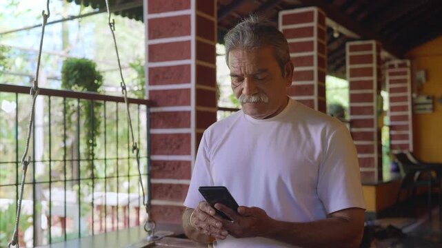 Daylight indoor close shot of a healthy, elderly Indian Asian man with grey hair sitting on a wooden swing on the veranda of a traditional house, using a mobile phone to type a text message