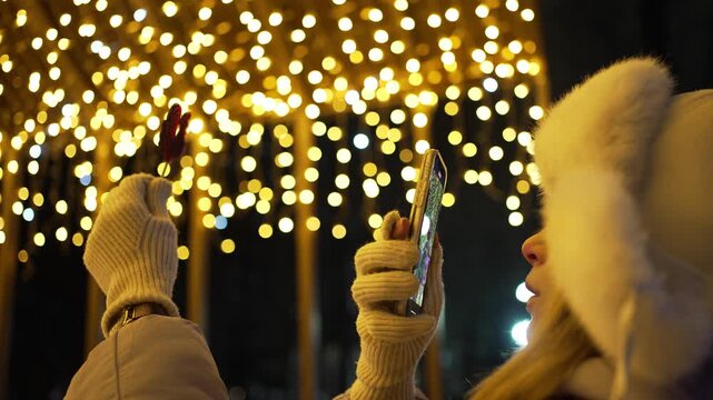 Young woman in warm winter clothes taking a photo with her mobile phone of a red rooster lollipop, with bright yellow festive bokeh lights illuminating the dark night