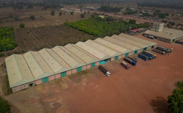 Aerial view of a large industrial warehouse with a sawtooth roof and parked trucks in a red dirt yard near the Abuja - Kaduna - Zaria Express Way in Kaduna, Kaduna, Nigeria.